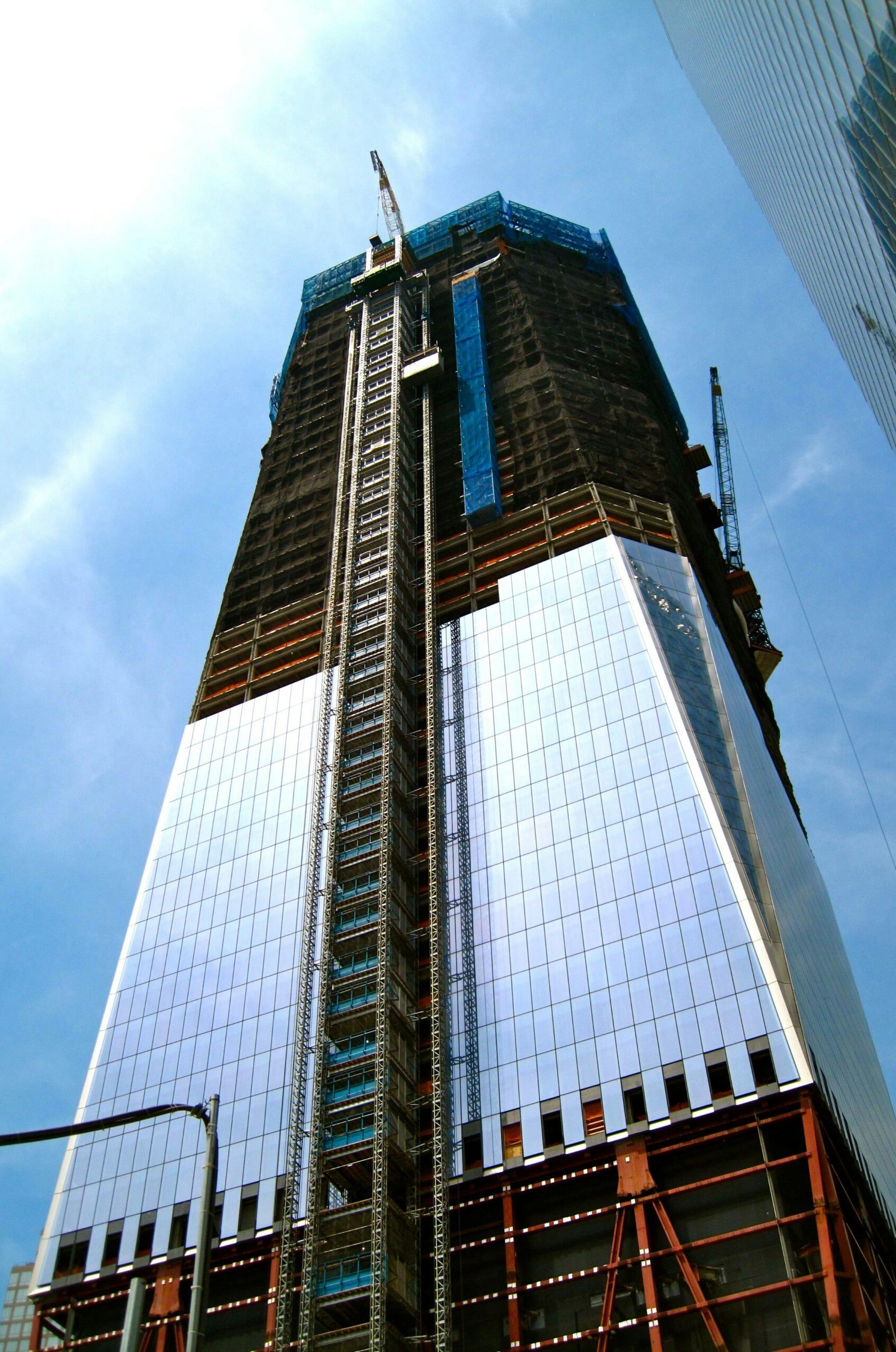 A towering modern skyscraper under construction with cranes and reflective glass against a clear blue sky.